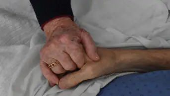 An older couple holds hands while husband is in palliative care