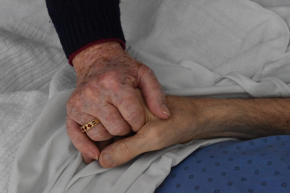 An older couple holds hands while husband is in palliative care