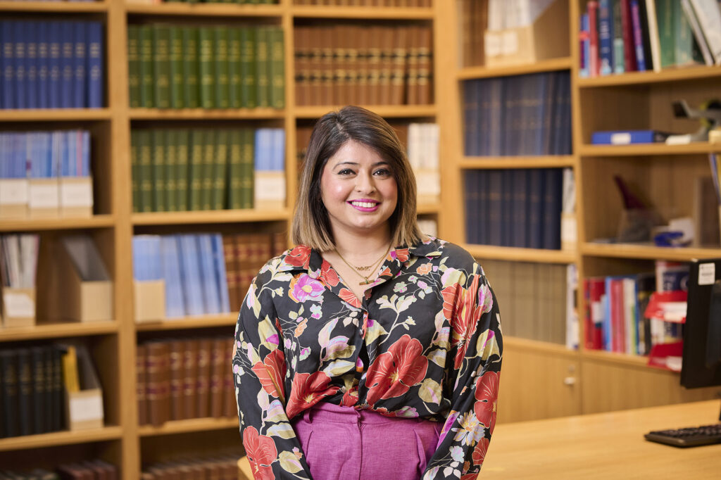 Amandeep smiles at the camera, wearing a floral shirt situated in a library