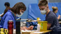 Meningitis vaccinations being given to students after the recent outbreak. woman with glasses on the left reading from a checklist to a male student on the right (both wearing masks). Surrounded by vaccination equipment