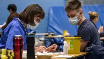 Meningitis vaccinations being given to students after the recent outbreak. woman with glasses on the left reading from a checklist to a male student on the right (both wearing masks). Surrounded by vaccination equipment