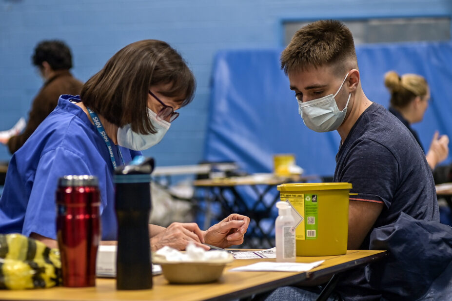 Meningitis vaccinations being given to students after the recent outbreak. woman with glasses on the left reading from a checklist to a male student on the right (both wearing masks). Surrounded by vaccination equipment