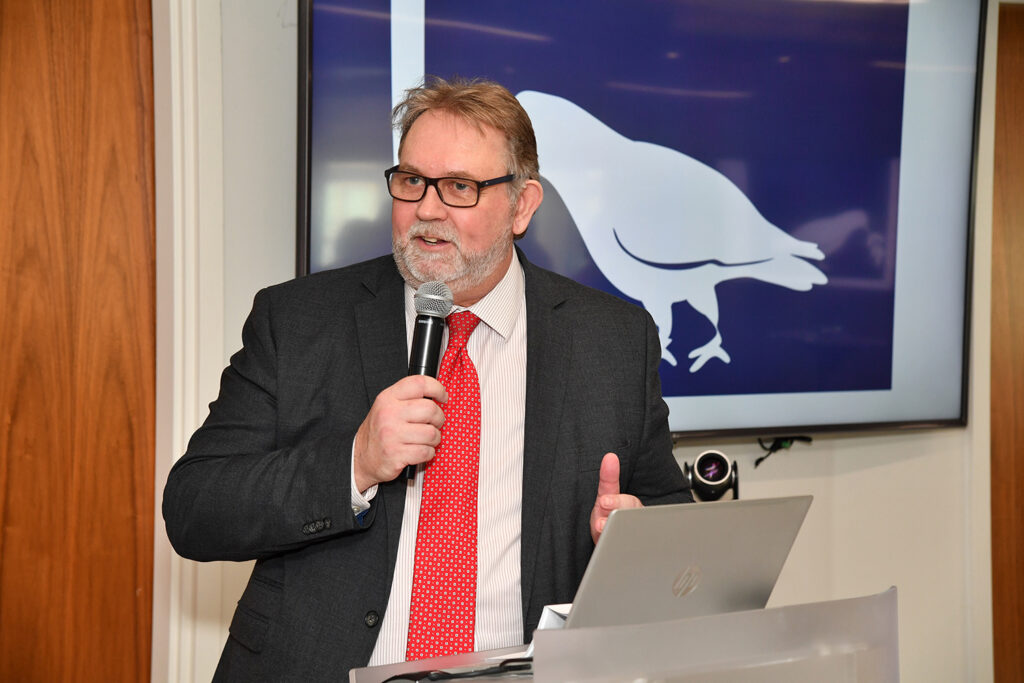 Paul wearing glasses, a suit and a red tie giving a talk at a podium in front of a screen