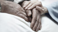 A loved one holds a patient's hand while they lay in a hospital bed