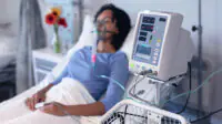 A woman sits in a hospital bed while on a ventilator