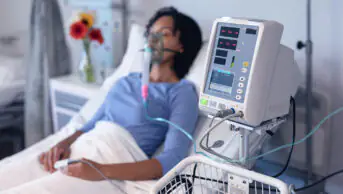 A woman sits in a hospital bed while on a ventilator