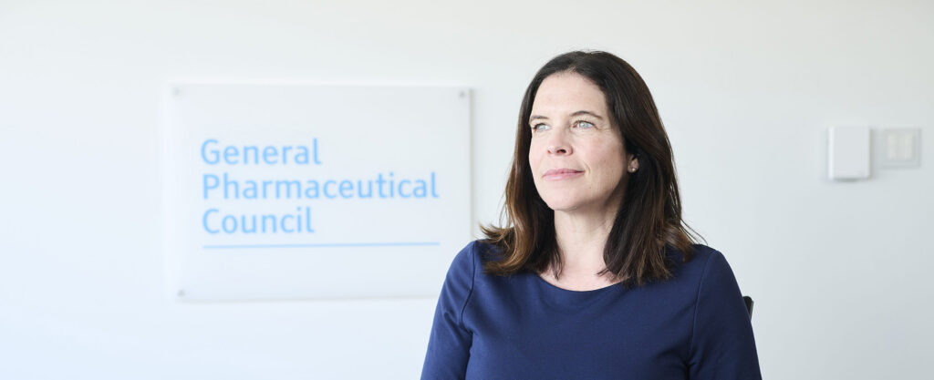 Kathie very faintly smiles while a dark shirt in front of a white wall with the General Pharmaceutical Council sign in the background