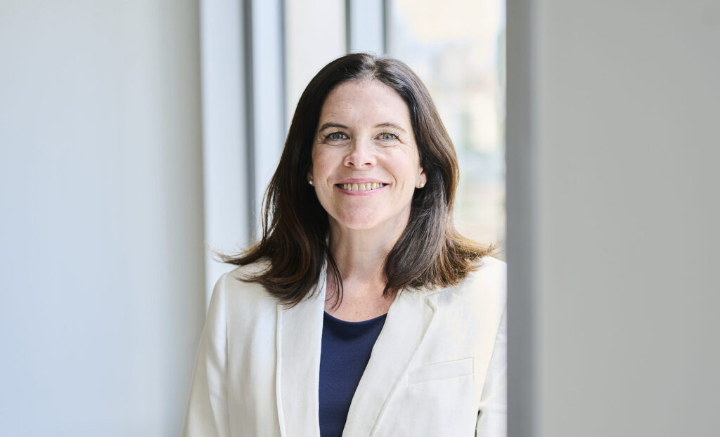 Kathie smiles while wearing a white blazer and dark shirt in front of a white wall and by a window shining light from the right