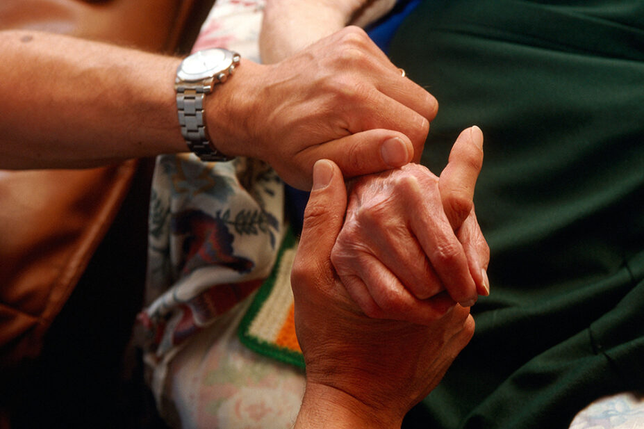 A loved one holds the hand of a patient in palliative care