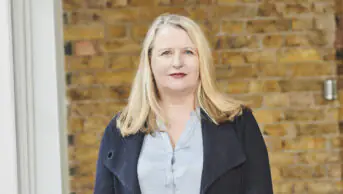 Janet stands in front of a brick wall, wearing a light blue shirt and dark blue blazer