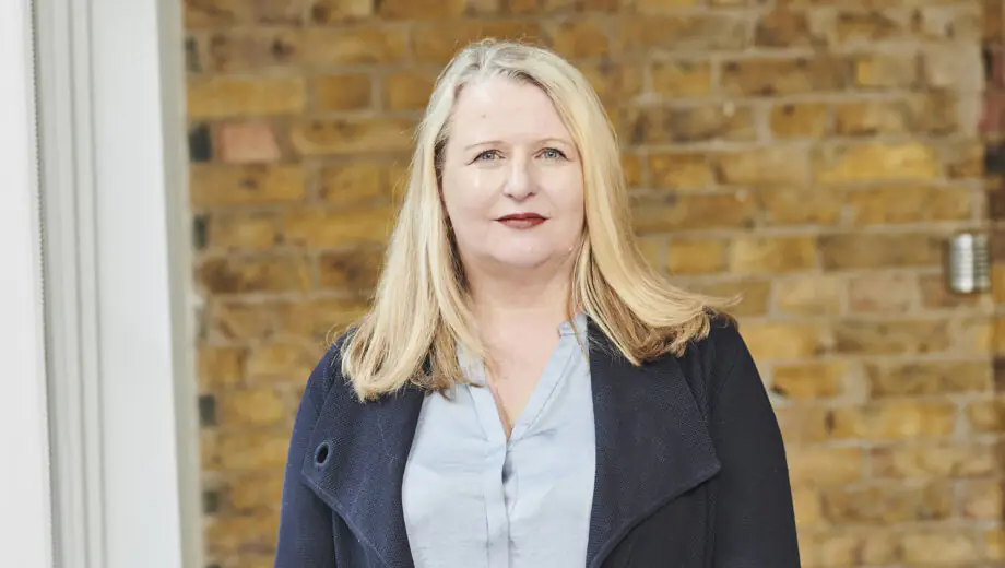 Janet stands in front of a brick wall, wearing a light blue shirt and dark blue blazer