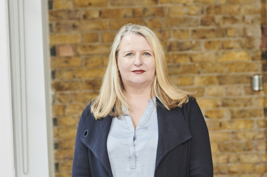 Janet stands in front of a brick wall, wearing a light blue shirt and dark blue blazer
