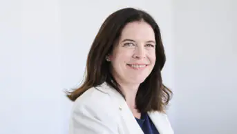 Kathie smiles while wearing a white blazer and dark shirt in front of a white wall