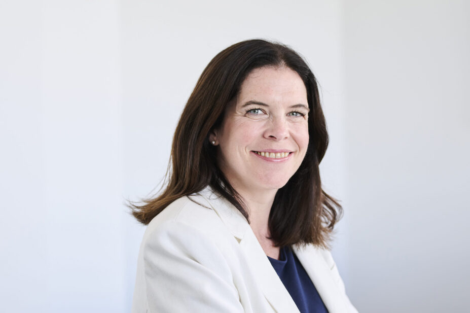 Kathie smiles while wearing a white blazer and dark shirt in front of a white wall