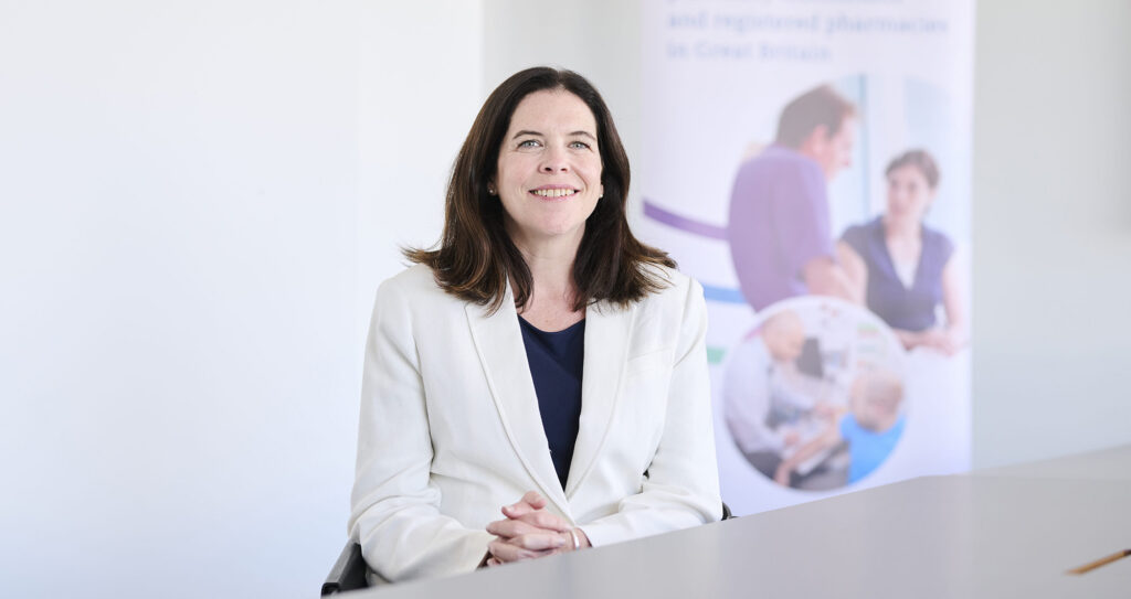 Kathie smiles while wearing a white blazer and dark shirt in front of a white wall and a GPhC banner