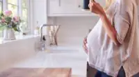 Close Up Of Pregnant Woman Standing By Kitchen Window Touching Stomach Relaxing With Hot Drink
