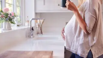 Close Up Of Pregnant Woman Standing By Kitchen Window Touching Stomach Relaxing With Hot Drink