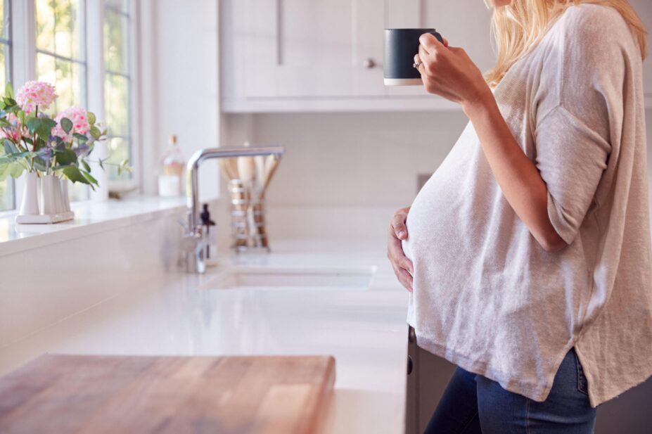 Close Up Of Pregnant Woman Standing By Kitchen Window Touching Stomach Relaxing With Hot Drink