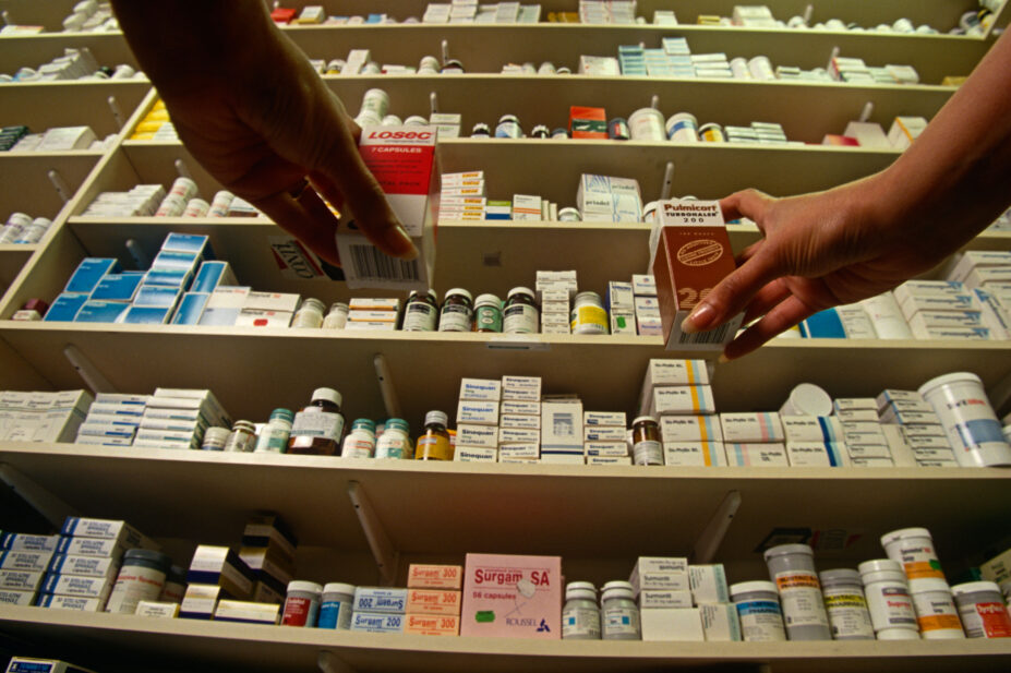 Customers reach for medicines on a shelf in a community pharmacy