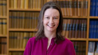 Geraldine standing against wooden bookshelf wearing dark pink shirt