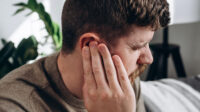 Close-up of unhealthy young caucasian man 30s touching ear, suffering from sudden throbbing