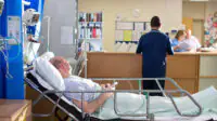 A hospital ward scene with a man in a bed, with clinicians in the background