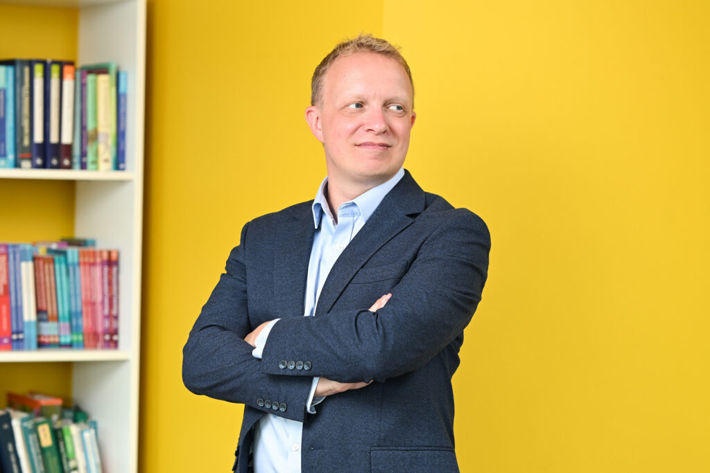 Alex looks serious and pensive looking to his left, wearing a light blue shirt and a dark blazer in front of a yellow wall with a bookcase