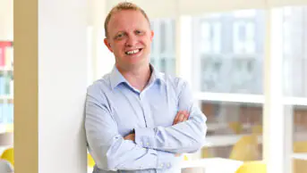 Alex smiles wearing a light blue shirt in front of a brightly lit room with chairs and books in the background