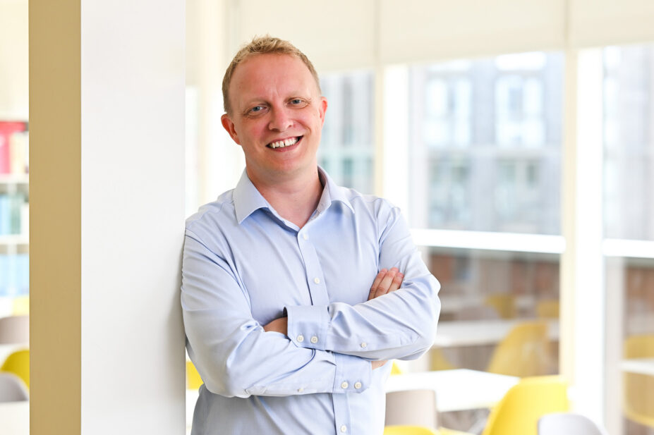 Alex smiles wearing a light blue shirt in front of a brightly lit room with chairs and books in the background