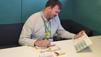 Michael sits at a desk smiling looking down at blisters packets and pill packets. The chair is dark blue and teal behind him