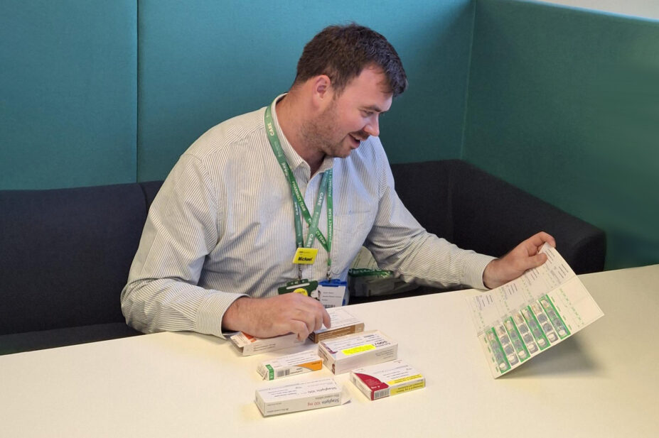 Michael sits at a desk smiling looking down at blisters packets and pill packets. The chair is dark blue and teal behind him