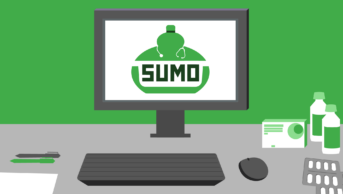 Green wall, grey desk, computer with SUMO logo on screen. On desk is keyboard, mouse, pens, pill packets and bottles