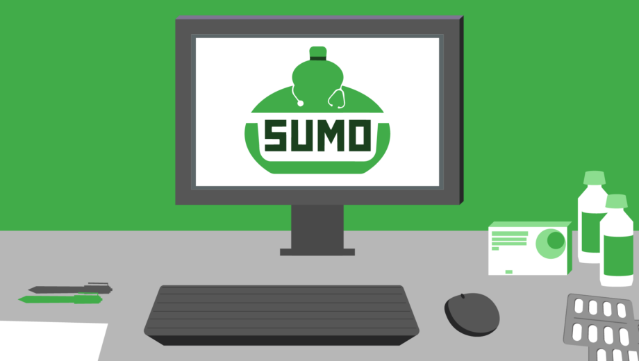 Green wall, grey desk, computer with SUMO logo on screen. On desk is keyboard, mouse, pens, pill packets and bottles