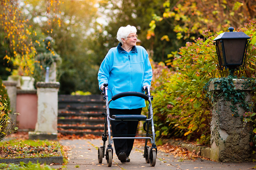 Older woman in blue jumper walks in leafy outside with walking aid