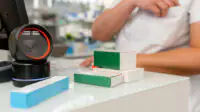 A pharmacist stands at the counter of the pharmacy with a barcode scanner and boxes of medicines