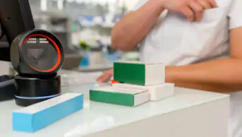 A pharmacist stands at the counter of the pharmacy with a barcode scanner and boxes of medicines