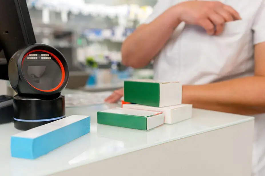 A pharmacist stands at the counter of the pharmacy with a barcode scanner and boxes of medicines