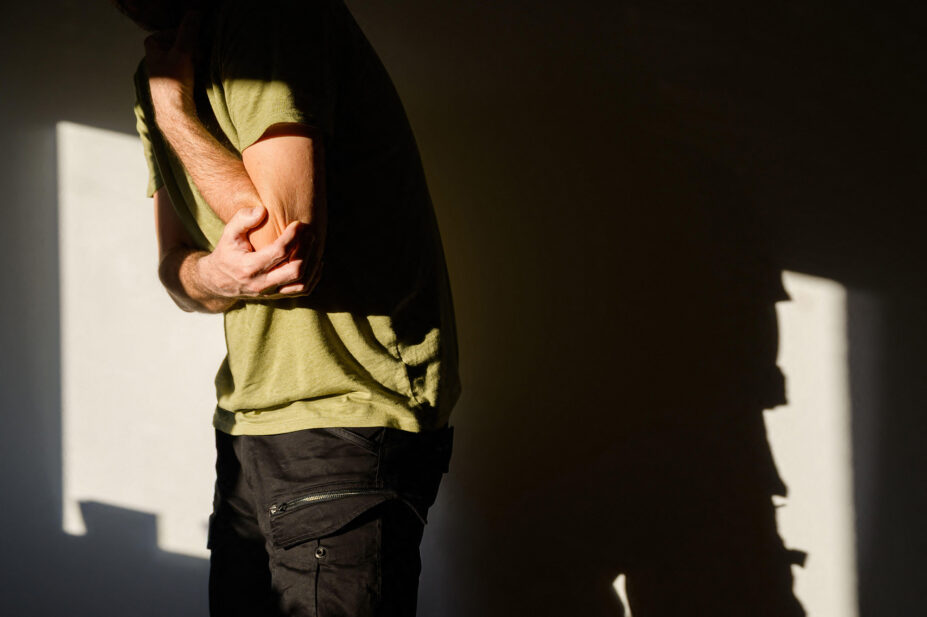 Middle-aged man wearing khaki colour t shirt gripping his elbow tightly with one hand in a gesture of acute joint pain or injury.