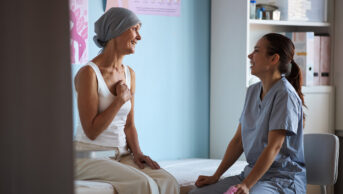 Middle aged Caucasian woman with headscarf sitting on examination table, smiling and talking with young adult female nurse, in medical clinic with breast cancer awareness posters visible