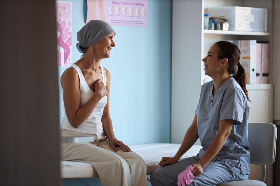 Middle aged Caucasian woman with headscarf sitting on examination table, smiling and talking with young adult female nurse, in medical clinic with breast cancer awareness posters visible