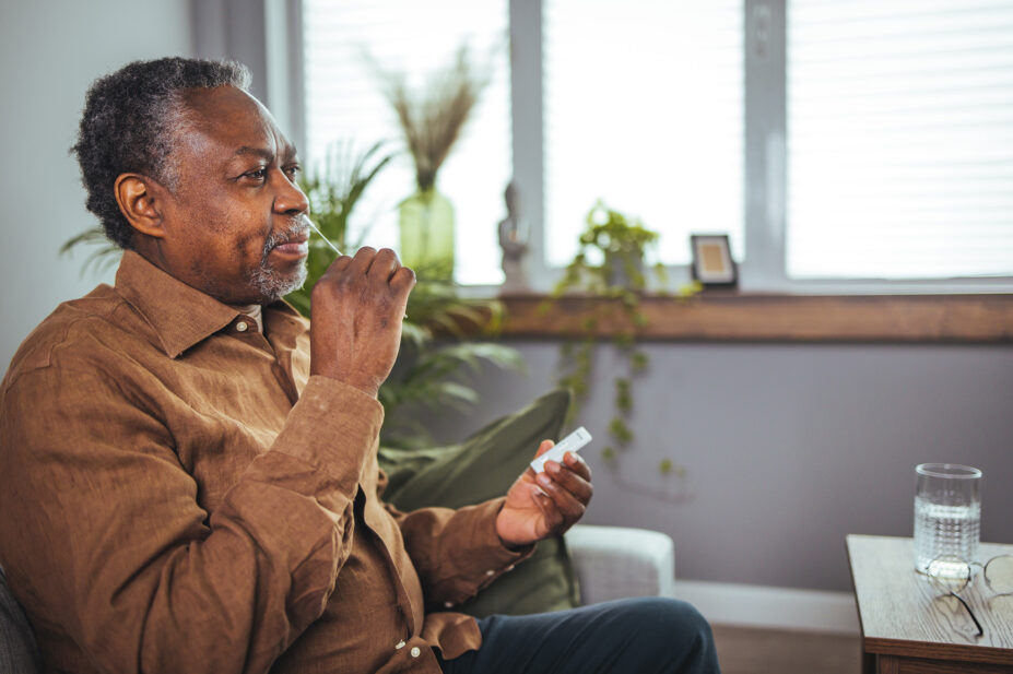 A senior black man in his 60s taking a Covid-19 lateral flow self-test at home, wearing an orange shirt and sat on a sofa