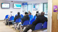 Person standing at reception while a busy waiting room in a health centre is busy and full with people sitting