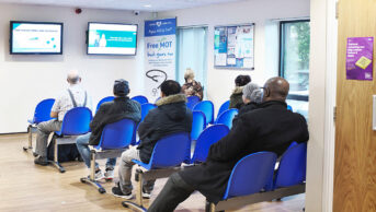 Person standing at reception while a busy waiting room in a health centre is busy and full with people sitting
