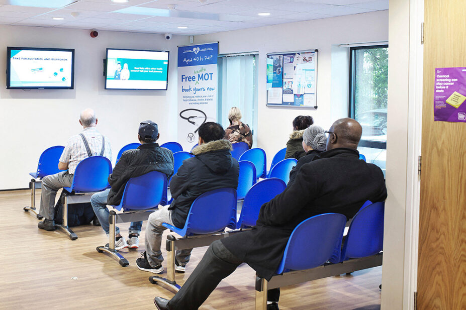 Person standing at reception while a busy waiting room in a health centre is busy and full with people sitting