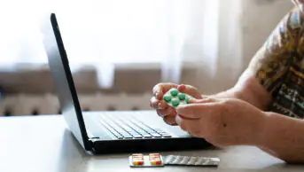 Elderly woman holding medicine, pills in hands, using laptop at home. Senior health, telemedicine. AI healthcare app, eHealth, e-pharmacy. Telehealth, remote video consultation, online therapy.