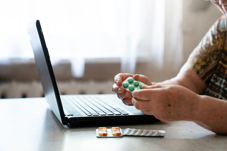 Elderly woman holding medicine, pills in hands, using laptop at home. Senior health, telemedicine. AI healthcare app, eHealth, e-pharmacy. Telehealth, remote video consultation, online therapy.