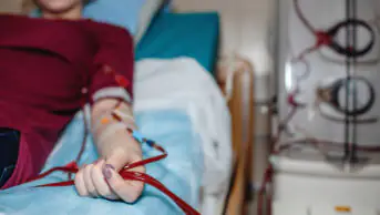 Woman in red top lies on hospital bed receiving dialysis from a machine on right