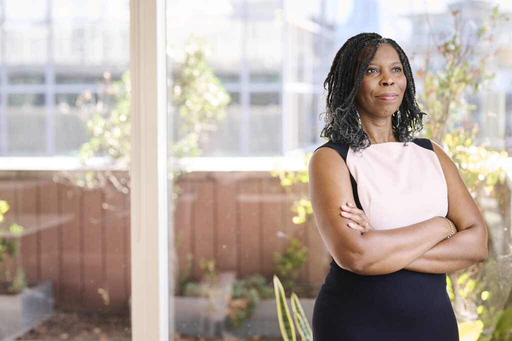Tase looks out a window with her arms crossed. The light from the window illuminates her face and arms. She wears a black and white dress. Behind her is a balcony and windows with plants