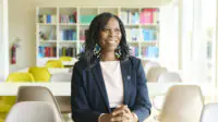 Tase smiles looking into the distance. She wears a white dress and navy blazer with a pin, blue and green earrings. Background is a sunny cafeteria with a bookcase visible in the back