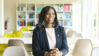 Tase smiles looking into the distance. She wears a white dress and navy blazer with a pin, blue and green earrings. Background is a sunny cafeteria with a bookcase visible in the back